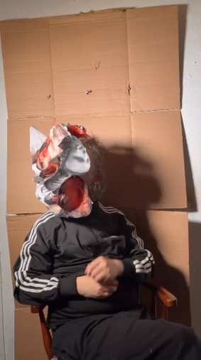 Crumpled Heads: John Robinson seated before a cardboard backdrop, wearing a mask built from crumpled prints of his own face.