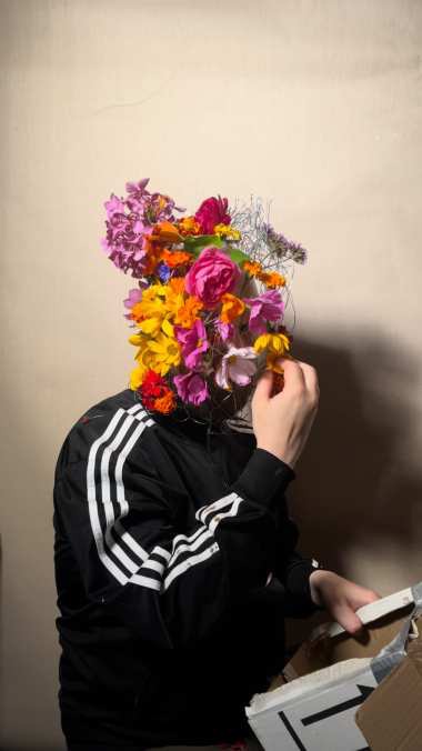 Flowerhead: John Robinson in a black track jacket, his head covered in fresh flowers threaded into a wire armature.