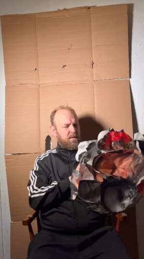 Still at 00:32 — Robinson seated before the cardboard backdrop, beginning to crumple printed images of his face.