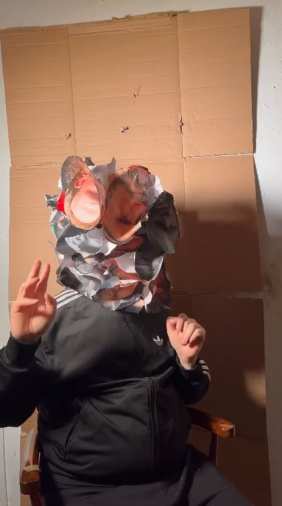 Still at 03:07 — the masked figure gesturing, face hidden among its damaged reproductions.