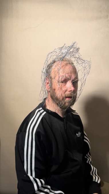 Still — Robinson wearing the bare wire armature over his head, before the flowers are threaded in.