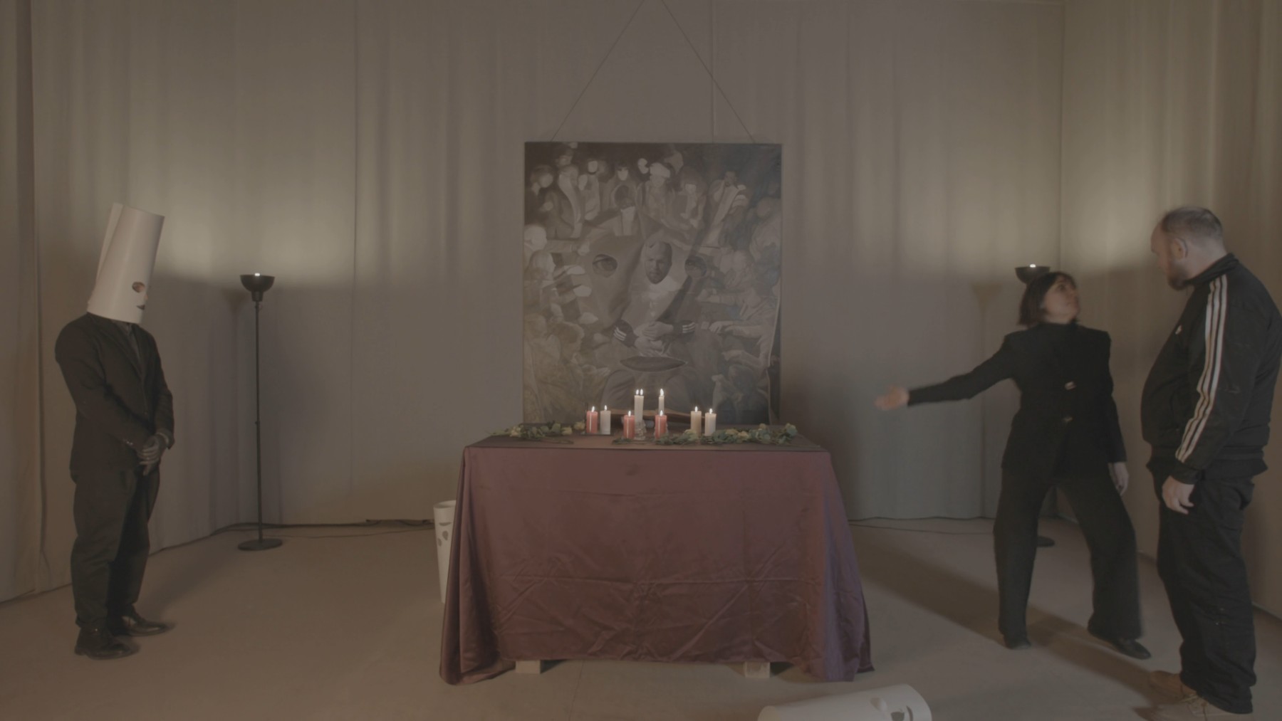 Still — the medium seated at the candlelit table, arms outstretched; three masked participants stand around her before a grisaille séance painting.